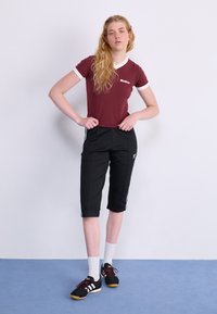 Bordeauxfarbenes Kurzarmhemd mit weißem Saum, schwarze knielange Hosen mit seitlichen Streifen, weiße Socken und schwarze Sportschuhe mit roten Akzenten.