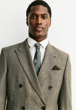 Man wearing a gray double-breasted suit with a white shirt and green floral-patterned tie, standing against a plain light background.