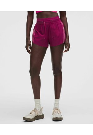 Kvinde iført lyserøde atletikshorts med side striber, beige sneakers og hvide sokker, stående mod en ensfarvet baggrund.