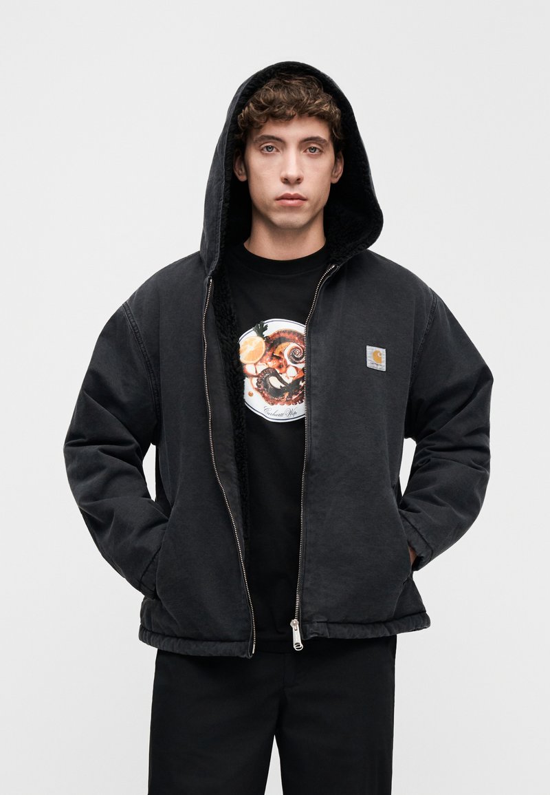 Sort hættejakke med lynlåslukning foran, fleecefor og et logopatch. Båret over en sort t-shirt med et cirkulært grafisk motiv.