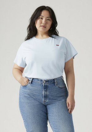 Kobieta z długimi ciemnymi włosami nosi jasnoniebieski t-shirt Levi's wpuszczony w wysoko taliowane niebieskie dżinsy na jednolitym szarym tle.
