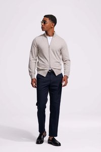 Lichtgrijze zip-up sweater over een wit shirt, gecombineerd met donkerblauwe broek en zwarte nette schoenen. Accessoires zijn groene zonnebrillen.