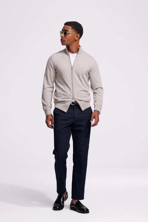 Lichtgrijze zip-up sweater over een wit shirt, gecombineerd met donkerblauwe broek en zwarte nette schoenen. Accessoires zijn groene zonnebrillen.