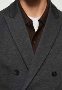 Blazer doppiopetto in tessuto grigio scuro con finitura testurizzata, caratterizzato da rever a punta, due bottoni e una camicia marrone sotto.