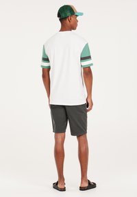 Casual mannenoutfit met een wit T-shirt met teal-accenten op de mouwen en gestreepte details, gecombineerd met donkere shorts en sandalen.