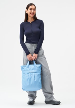 Frau in marineblauem Langarmshirt und grauer Hose, die eine hellblaue Tragetasche hält, steht lächelnd vor weißem Hintergrund.