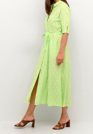 Frau trägt ein hellgrünes, gemustertes Midikleid mit Taillengürtel und braunen Sandalen mit Absatz, steht vor einem weißen Hintergrund.