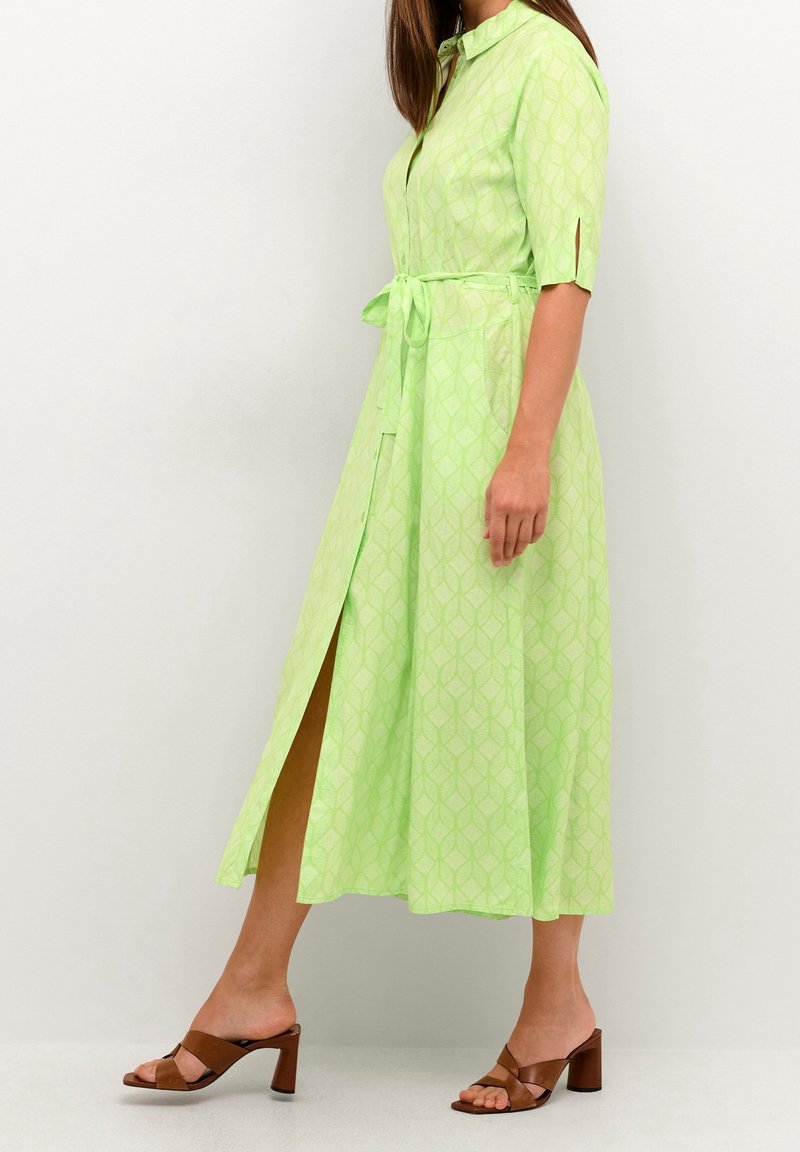 Femme portant une robe chemise midi à motifs vert clair avec une ceinture à la taille et des sandales à talons marron, debout devant un fond blanc.