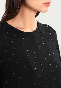 Garbamento nero con scollatura rotonda, caratterizzato da una trama liscia e da un motivo di piccoli punti riflettenti color oro rosa distribuiti uniformemente.