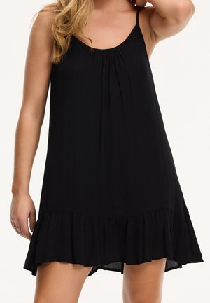 Kobieta ubrana w czarną, bezrękawnikową sukienkę z cienkimi ramiączkami i falbaną u dołu, stojąca z jedną uniesioną ręką, a drugą swobodnie opuszczoną.