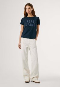 T-shirt in cotone navy con grafica "PEPE JEANS" in colori chiari, abbinata a pantaloni larghi bianchi e scarpe beige. Design semplice e casual.