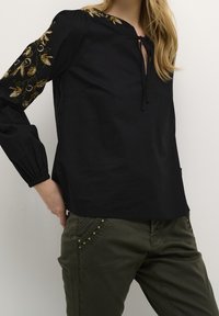 Blouse noire avec une patte de boutonnage et un nœud au cou, ornée de broderies florales dorées sur les manches. Associée à un pantalon vert olive.