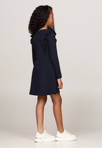 Abito navy a maniche lunghe con accenti a volant lungo la schiena, dotato di una gonna a campana; abbinato a sneakers bianche. Texture liscia.