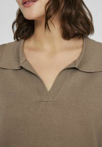 Jersey marrón con un cuello tipo polo y un corte en forma de V. La textura es suave con detalles acanalados a lo largo del cuello.