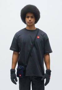 Sort t-shirt med korte ærmer og rødt logo, sorte handsker og en lille navy crossbody taske, båret af en person foran en ensfarvet baggrund.