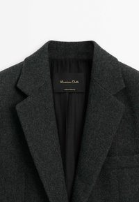 Harmaa villasekoitebleiseri, jossa on viisto kaulus ja musta vuori. Varustettu etiketillä, jossa lukee "Massimo Dutti" kultaisella tekstillä. Siisti muotoilu, ei näkyviä nappeja.