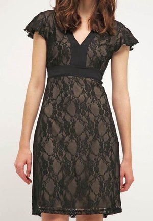Frau in einem schwarzen Spitzenkleid mit kurzen Rüschenärmeln und V-Ausschnitt über einer hautfarbenen Unterlage, stehend vor einem schlichten Hintergrund.