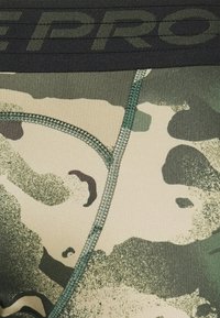 Gros plan sur un tissu camouflage vert et beige avec des coutures visibles et un texte partiel sombre "PRO" sur une bande noire.