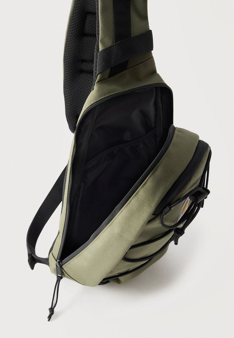 Grüne Crossbody-Tasche mit schwarzem Reißverschluss geöffnet, zeigt schwarzes Innenfutter und gepolsterten Rückengurt mit Mesh-Details.