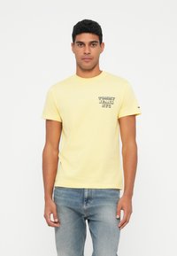 Geltona medvilninė marškinėliai su apvaliu kaklu, kuriuose yra maža "TOMMY JEANS NYC" grafika ant krūtinės. Sujungti su šviesiai mėlynais džinsais.