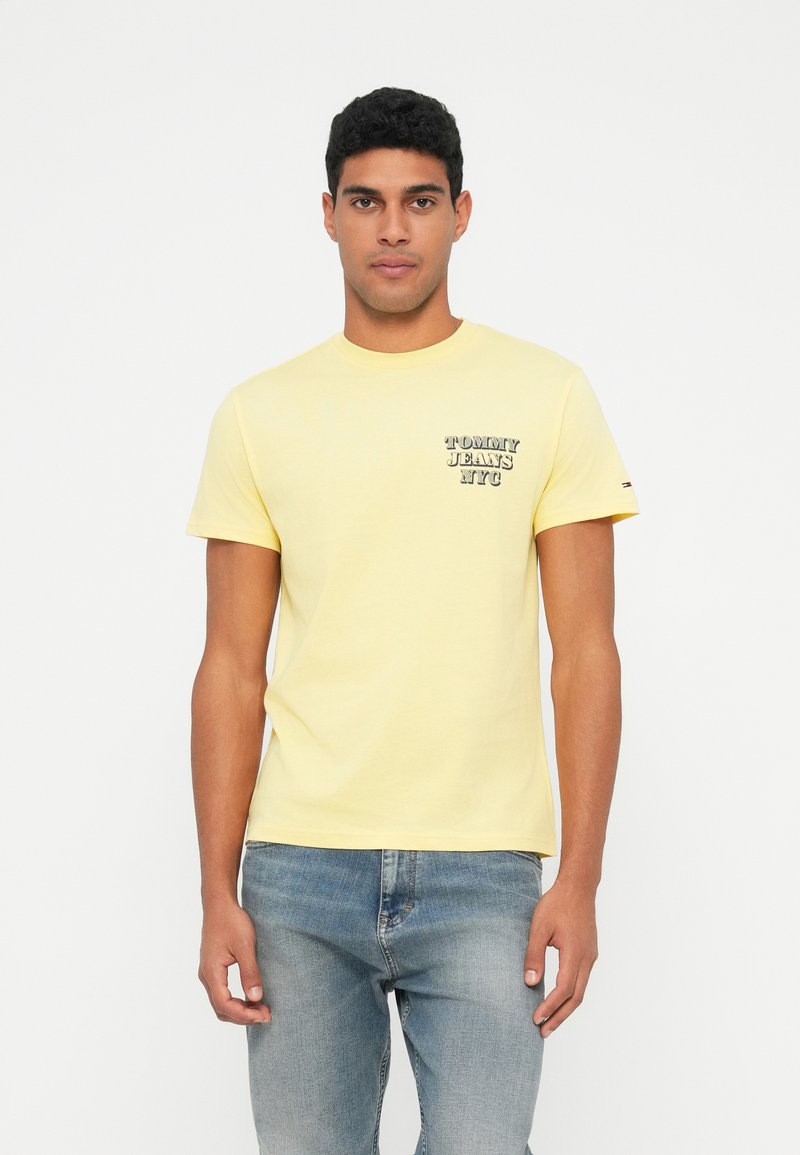 Żółta bawełniana koszulka z okrągłym dekoltem, z małym nadrukiem "TOMMY JEANS NYC" na piersi. W zestawie z jasnoniebieskimi dżinsami.