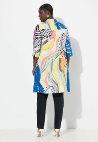 Giacca in stile kimono colorata e decorata, con colletto, caratterizzata da design astratti blu, arancio e nero; abbinata a pantaloni neri e tacchi.