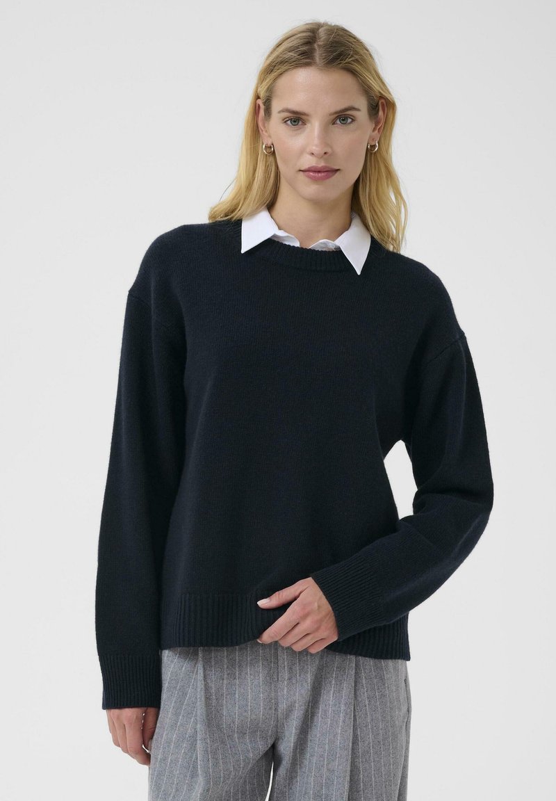Dark navy knit sweater with a round neckline, dropped shoulders, and ribbed cuffs, worn over a white collared shirt, paired with gray striped trousers.