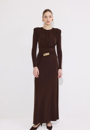 Femme debout contre un mur blanc, portant une robe marron à manches longues avec un détail boucle dorée et des talons marron assortis.