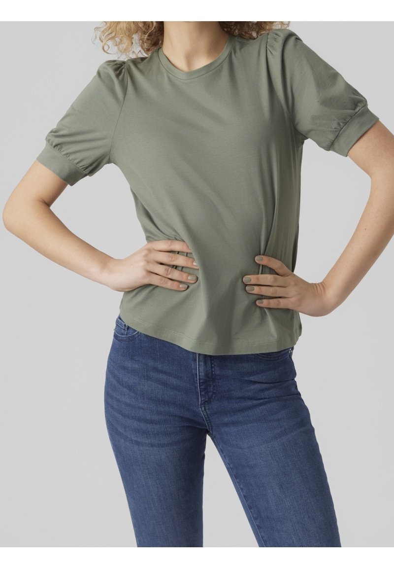 Personne aux cheveux bouclés portant une chemise verte à manches courtes avec des épaules bouffantes et un jean bleu, les mains sur les hanches, sur un fond uni.