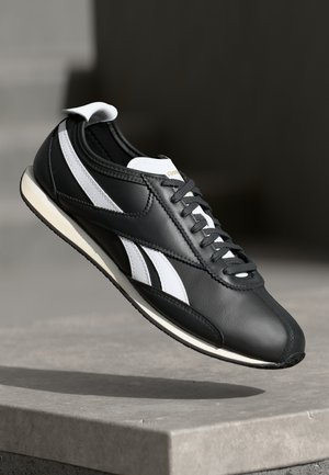 Baskets Reebok noir et blanc avec logo et lacets blancs, positionnées en l'air au-dessus d'une surface en béton sur un fond sombre flou.