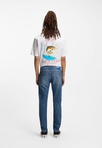 Λευκό T-shirt με κοντά μανίκια, με γραφικό ψαρέματος και κόκκινο κείμενο στην πίσω πλευρά, σε συνδυασμό με μπλε τζιν παντελόνι και μαύρα αθλητικά παπούτσια με λευκές λεπτομέρειες.