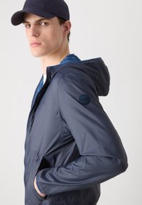 Marineblaue Kapuzenjacke mit glänzender Oberfläche, Reißverschluss vorne und seitlichen Taschen. Ausgestattet mit einem runden Markenpatch auf dem linken Ärmel.