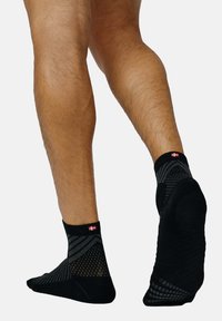 Calcetines negros hasta el tobillo con un diseño estructurado, que presenta un patrón de malla y un logo en rojo y blanco en la parte superior. Textura suave, estilo de corte bajo.