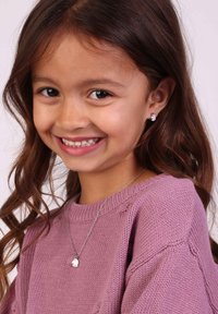 Zilveren ketting met een klein bedeltje, gedragen over een zachtroze gebreide trui. Kind met lang, golvend bruin haar en een vriendelijke glimlach.