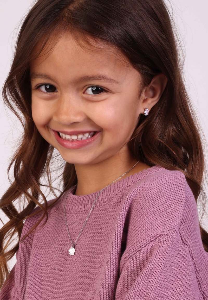 Zilveren ketting met een klein bedeltje, gedragen over een zachtroze gebreide trui. Kind met lang, golvend bruin haar en een vriendelijke glimlach.