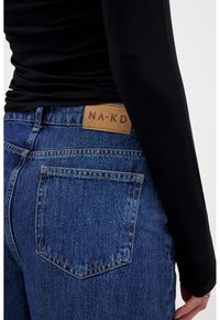Vaqueros de denim en azul medio con cintura alta, que presentan una etiqueta de cuero de NA-KD y un bolsillo trasero. La tela tiene un acabado texturizado.