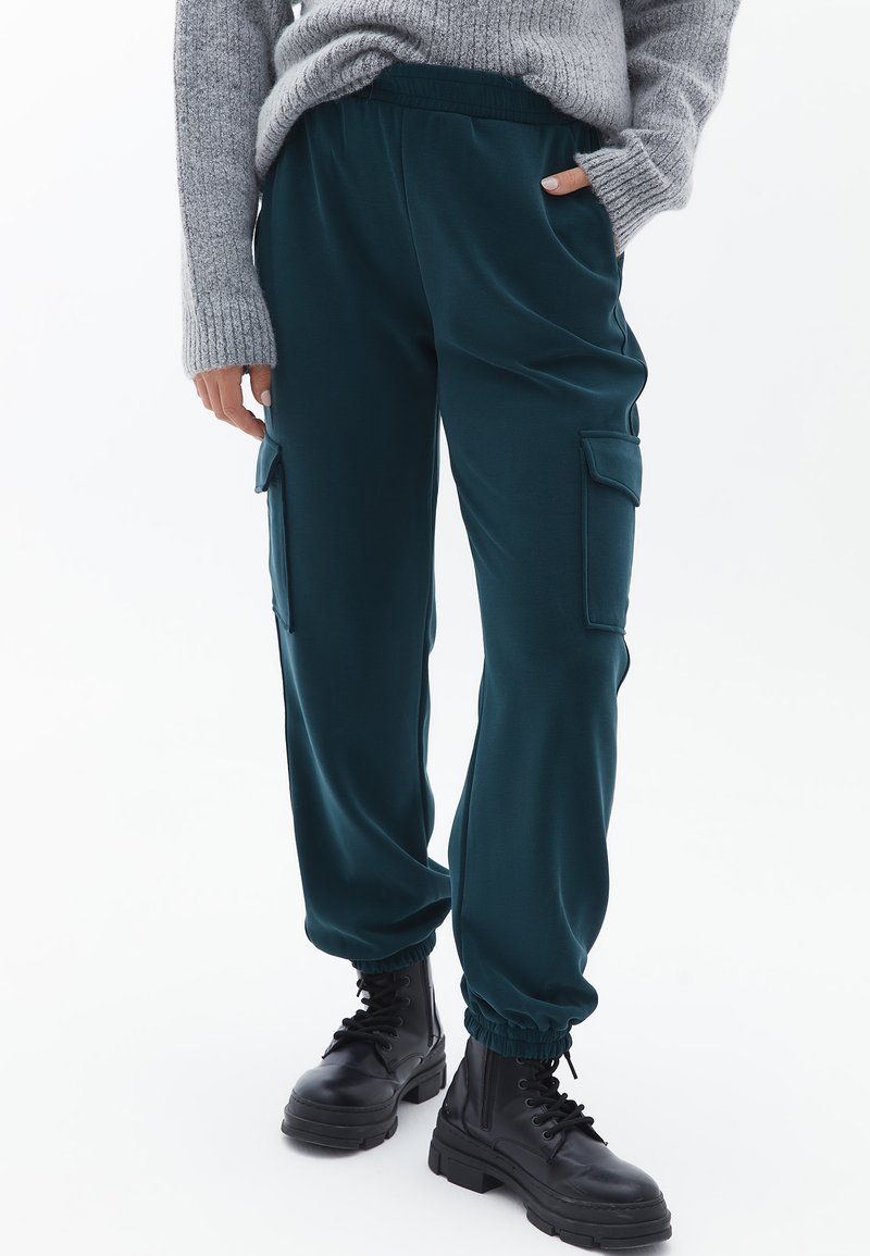 Pantaloni jogger teal con elastiche in vita e sui polsini, dotati di tasche laterali e una texture liscia, abbinati a stivaletti neri chunky.