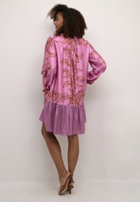 Ružové šaty s kvetinovým vzorom a volánovým lemom. Majú dlhé rukávy a svetlú, lesklú textúru látky. Dĺžka po stred stehna.