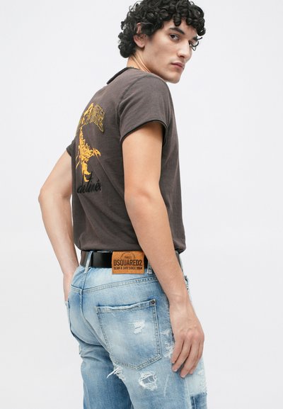 T-shirt graphique marron avec un design de scorpion et un jean bleu clair usé, accompagné d'une ceinture noire avec un patch logo beige.