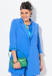 Sininen bleiseri, jonka hihat on käännetty, kerroksittain teal-värisen paidan päällä. Pitää käsissään pientä vihreää laukku, jonka pinnassa on tekstuuria, ja hopeiset korut ovat näkyvissä.