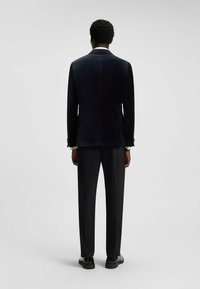 Blazer in velluto navy con spalle strutturate, forma aderente e texture liscia, abbinato a pantaloni neri su misura e scarpe nere lucide.