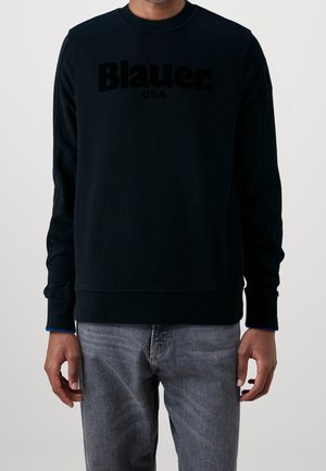 Man wearing a black long-sleeve sweatshirt with "Blauer USA" text on the chest and gray jeans against a plain background.