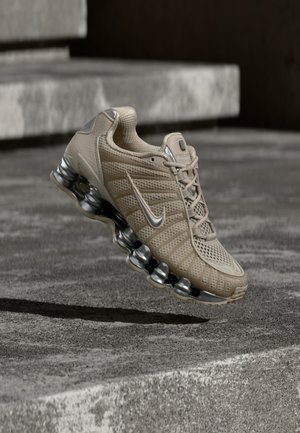 Chaussure de sport beige avec des détails argentés et une semelle à coussin d'air, suspendue au-dessus d'une surface en béton avec une ombre en dessous.