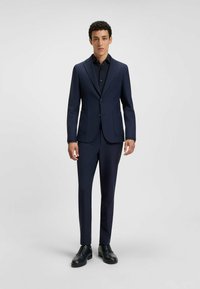 Abito navy realizzato in tessuto liscio. Giacca aderente con due bottoni e tasca, abbinata a pantaloni coordinati e scarpe nere.