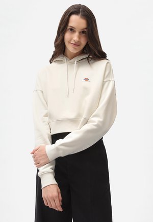 Cropped cream hoodie with drawstrings and a small logo. Soft texture, relaxed fit. Model wearing black pants against a plain background.