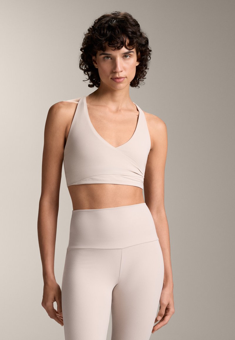 Bej színű sportmellény V-nyakú és átkötős dizájnnal, magas derekú, varrás nélküli leggings-szel párosítva, megegyező színben. Simás, rugalmas anyag.