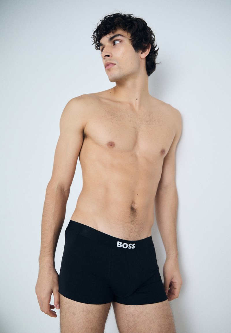 Schwarze Boxershorts aus einem glatten Material mit einem breiten Gummibund, der den Markennamen "BOSS" in weiß zeigt. Glatte Textur, enganliegendes Design, minimalistischer Stil.