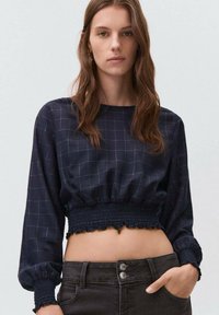 Avskuren marinblå blus med rutmönster, insamlad elastisk nederkant och långa ärmar med volanger, som är ihop med mörka jeans.