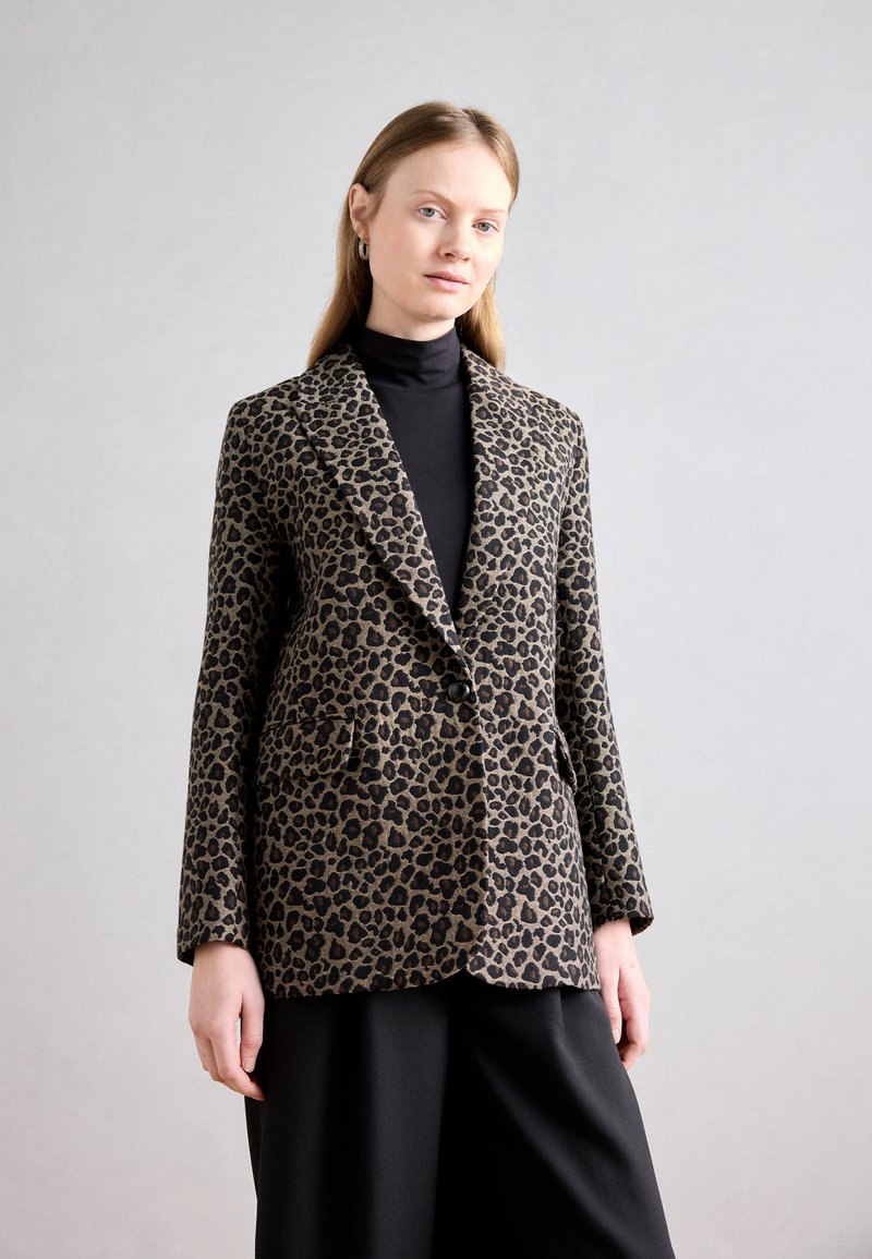 Leopardenmuster Blazer in Beige und Schwarz, mit breitem Revers, zwei vorderen Taschen und einem einzelnen Knopfverschluss. Getragen über einem schwarzen Rollkragenpullover.