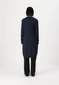 Largo cárdigan azul marino hecho de material de punto suave, con un corte recto, puños y dobladillo acanalados, con un detalle mínimo, usado sobre pantalones negros.